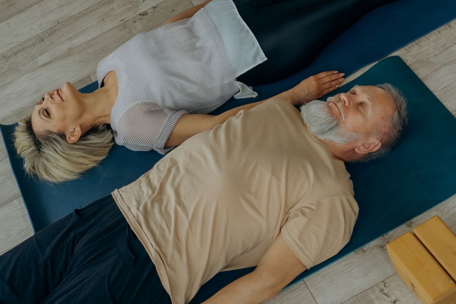 Two people resting side by side on a yoga mat with eyes closed, breathing together