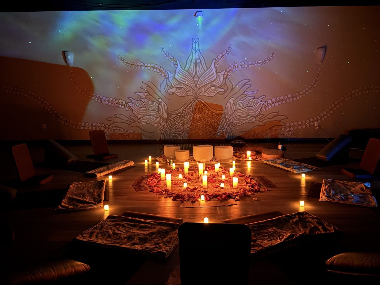 A candlelit sound healing circle with crystal bowls, rose petals and cushions arranged in a ring